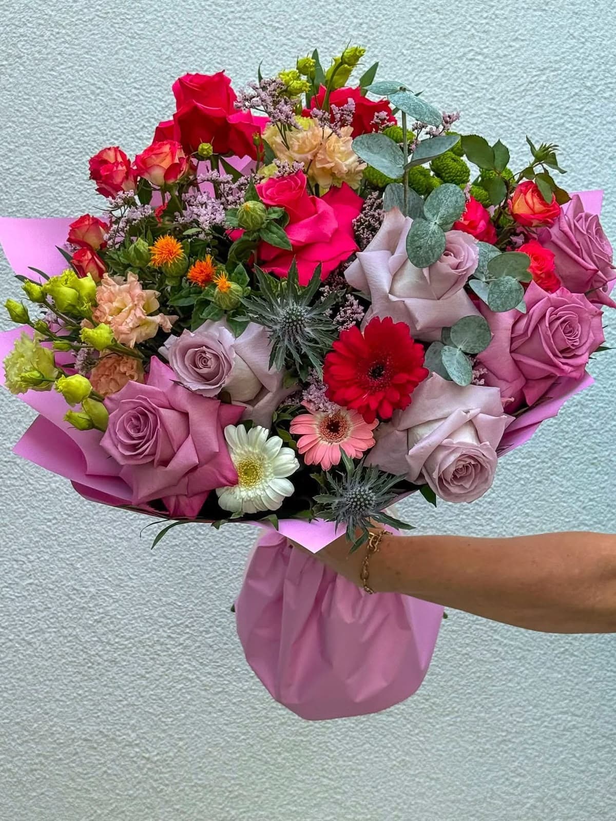 Buchet colorat cu trandafiri ciclam și mov, gerbera și scaieți albaștri, ambalat în hârtie roz