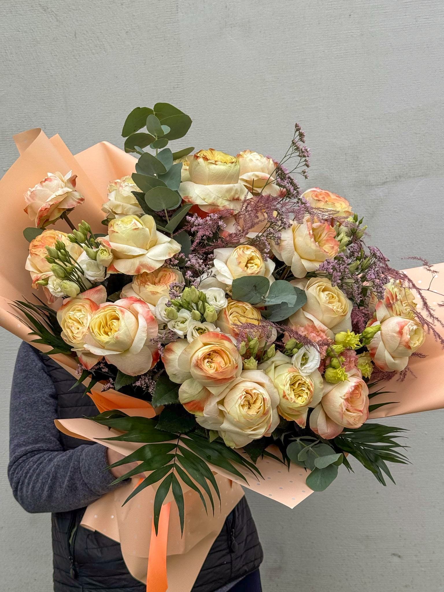 Buchet elegant cu trandafiri crem și piersică, completat cu eucalipt, flori decorative mov și frunziș verde. Un aranjament floral premium, în ambalaj pastel, ideal pentru aniversări, ocazii speciale și cadouri rafinate.