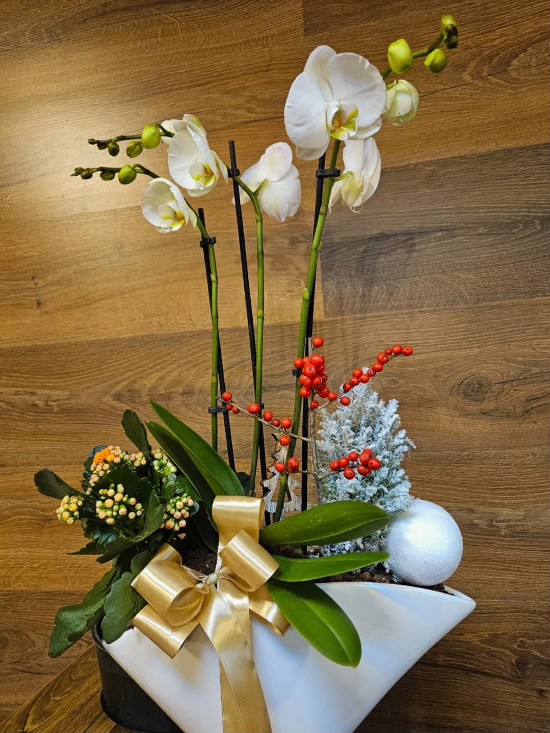Aranjament floral elegant cu orhidee albe, kalanchoe și ilex cu bobițe roșii, decorat cu elemente decorative de iarnă și panglică aurie, în vas ceramic alb. Ideal pentru cadouri festive și decorarea locuinței de Crăciun.